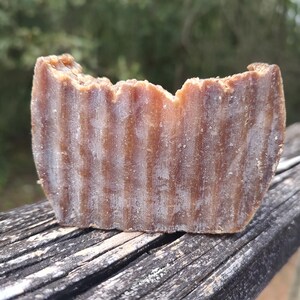 Puede incluir: Una pastilla rectangular de jabón marrón con una superficie texturizada y acanalada. El jabón tiene un borde superior ligeramente irregular y parece haber sido usado. Descansa sobre una superficie de madera envejecida.