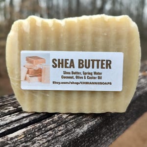 May include: A bar of shea butter soap with a textured surface. The white label reads "SHEA BUTTER" and lists ingredients: Shea Butter, Spring Water, Coconut, Olive & Castor Oil. The soap is rectangular.