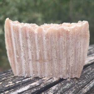 Puede incluir: Una pastilla rectangular de jabón hecho a mano con una superficie texturizada y estriada. El jabón es de color beige claro con vetas blancas y un borde superior ligeramente irregular. El jabón está sobre una superficie de madera envejecida.