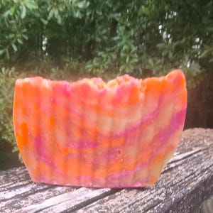 Puede incluir: Una barra rectangular de jabón hecho a mano con un vibrante patrón de remolino de tonos naranja, rosa y amarillo. El jabón descansa sobre una superficie de madera desgastada, con un fondo verde borroso de follaje.