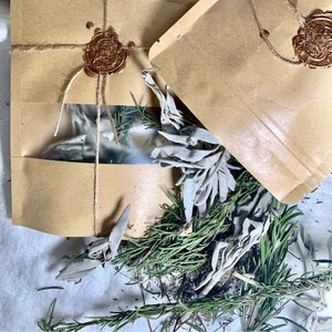 May include: Two brown paper pouches sealed with a gold wax stamp and twine. One pouch has a clear window. Bundles of green rosemary and white sage are arranged around the pouches. A silver religious medallion is visible in the background.