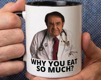 May include: White ceramic mug with a black interior and handle, featuring an image of a man in a white coat and a red tie. The text "WHY YOU EAT SO MUCH?" is printed below the image. The mug is held by a person.