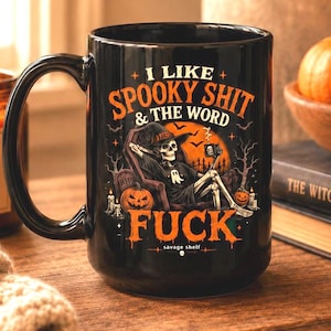 May include: A black ceramic mug featuring a graphic of a skeleton sitting in a chair, surrounded by pumpkins, trees, and tombstones. The text reads I LIKE SPOOKY SHIT & THE WORD FUCK. The design includes orange and white accents on the black surface.
