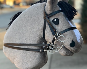 May include: A plush gray stick horse with black mane and bridle. The horse has a white stripe on its face and black eyes. The bridle is made of black leather and has silver-colored metal accents. The stick is brown.
