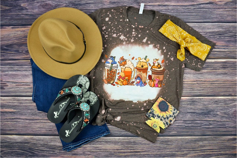 A t-shirt with a winnie the pooh design pressed onto it. A pair of jeans and flipflops is beside the shirt.
