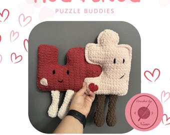 May include: Two crocheted puzzle piece plushies, one red and one pink, with smiling faces and heart details. The plushies are holding hands and have brown legs. The text "HUG & SNUG PUZZLE BUDDIES" is at the top of the image. The text "Crochet by Nour" is in a circle at the bottom right of the image.