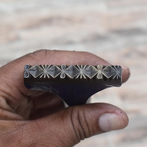 Könnte beinhalten: Ein dunkles Metallstempelwerkzeug mit einem rechteckigen Kopf, der ein sich wiederholendes geometrisches Muster aus sternartigen Formen und Rautenmotiven auf der Oberfläche aufweist, entworfen für Abdrücke in Leder oder Bastelmaterialien.