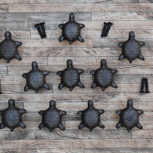 Op de afbeelding: Tien zwarte gietijzeren schildpad-ladegrepen met schroeven. De grepen zijn symmetrisch gerangschikt op een houten oppervlak.