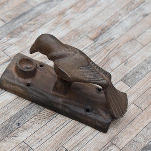 Puede incluir: Un aldaba de puerta con forma de pájaro de hierro fundido con un acabado bronce envejecido. El pájaro está posado sobre una base rectangular con un cuenco pequeño y agujeros de montaje. La aldaba está diseñada para montarse en una puerta.