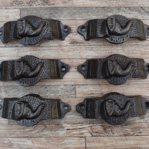 Può includere: Sei tiretti per cassetti in ghisa nera con un design a forma di elefante. Ogni tiretto ha una superficie testurizzata e una testa di elefante in rilievo con una proboscide.