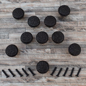 May include: Ten black metal cabinet knobs with a decorative floral design, arranged in a circle on a wood surface.  The knobs are accompanied by ten black screws.
