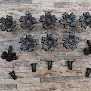 Op de afbeelding: Twaalf zwarte gietijzeren haken in de vorm van een bloem met een decoratief ontwerp. De haken zijn gerangschikt in twee rijen van zes. Er zijn ook twaalf schroeven inbegrepen.