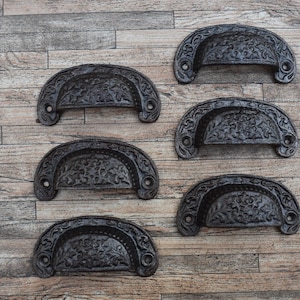 May include: Six antique-style, cast iron drawer pulls with a dark, distressed finish. Each pull has a semi-circular shape with ornate floral detailing. The pulls are arranged on a wooden surface.