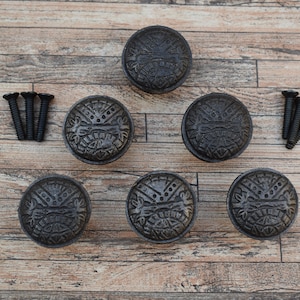 Può includere: Set di sei pomelli per armadi rotondi grigio scuro con un design in rilievo, insieme a due set di tre viti nere. I pomelli presentano un motivo dettagliato e strutturato. L'hardware è esposto su una superficie di legno marrone chiaro.