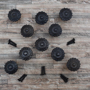 Könnte beinhalten: Zehn schwarze dekorative Schrankknöpfe mit floralem Design und mehrere schwarze Schrauben sind auf einer hellbraunen Holzoberfläche angeordnet. Die Knöpfe sind aus Metall.