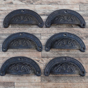 Könnte beinhalten: Sechs schwarze Schubladengriffe aus Gusseisen mit einem verzierten Design. Die Griffe sind in zwei Reihen zu je drei auf einem Holzgrund angeordnet.