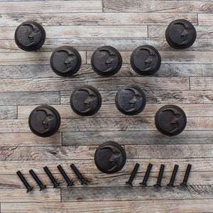 May include: Ten black metal cabinet knobs with a crescent moon design and a face in the centre. The knobs are arranged in a triangle shape on a wooden surface. Ten black screws are also included.