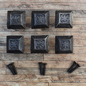 May include: Six black cast iron square drawer pulls with a decorative floral design. The pulls are arranged in two rows of three. The pulls are on a wooden surface.