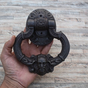 Peut inclure: Un heurtoir de porte en fonte noire avec un design détaillé représentant un visage grotesque et des volutes ornées.