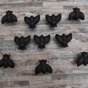 May include: Nine black cast iron bee-shaped drawer pulls on a wooden background.