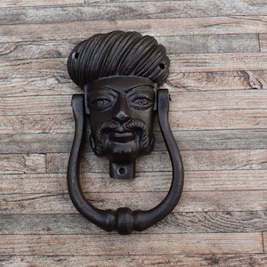 May include: A cast iron door knocker featuring a detailed face with a mustache and turban. The knocker is dark brown and has a ring-shaped handle. The door knocker is mounted on a wooden surface.