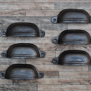 May include: Six black cast iron drawer pulls with a rounded, slightly curved shape. The pulls are arranged in two rows of three on a wood background.