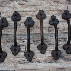 May include: Six black metal drawer pulls with a clover-shaped design. The pulls are arranged in a row on a wood surface.