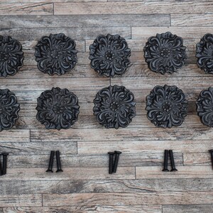 Könnte beinhalten: Ein Set aus zehn schwarzen dekorativen Schrankknöpfen mit floralem Design, angeordnet in zwei Reihen. Die Knöpfe sind aus dunklem Metall gefertigt und werden mit passenden Schrauben geliefert.