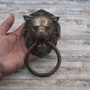 Peut inclure: Un heurtoir de porte en laiton en forme de tête de lion avec une poignée circulaire. Le heurtoir est détaillé avec une crinière texturée et un visage de lion réaliste.