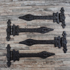 May include: Four black metal hinges with ornate designs, arranged in a row on a woodgrain background.