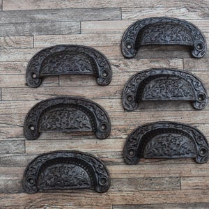 May include: Six black cast iron drawer pulls with an ornate floral design. The pulls are arranged in two rows of three on a wood background.