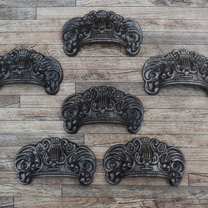 May include: Six black cast iron drawer pulls with ornate designs, including a lyre, flowers, and scrolls. The pulls are arranged in two rows of three on a wooden background.