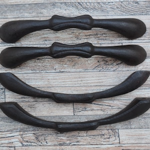 Può includere: Quattro maniglie per cassetti in ghisa nera con un design decorativo a forma di fiocco. Le maniglie sono disposte in fila su una superficie in legno.