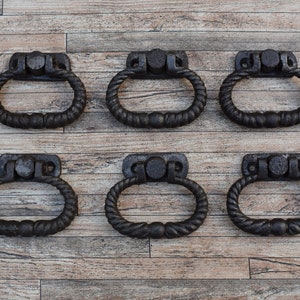 May include: Six black cast iron drawer pulls with a rope design. The pulls have an oval shape and are mounted on a rectangular plate.