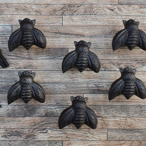 以下が含まれることがあります： 木製の表面に、テクスチャ加工が施された黒い鋳鉄製の蜂の形をした引き出しの取っ手が6つ並んでいます。取っ手は2列に3つずつ配置されています。