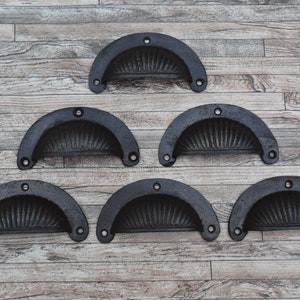 May include: Six black cast iron drawer pulls with a decorative sunburst design. The pulls are arranged in a triangular pattern on a wooden background.