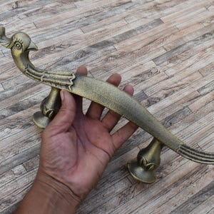 Puede incluir: Un tirador de puerta de metal en tono latón con forma de ave estilizada, cuerpo largo y curvado y cabeza con cresta. El tirador presenta detalles de alas texturizadas y dos bases de montaje circulares bajo el cuerpo del ave.