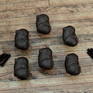 Peut inclure: Six boutons de meuble en fonte de couleur marron foncé avec un motif animalier détaillé, disposés sur une surface en bois. Sont également inclus deux jeux de vis noires pour l'installation. Ces boutons décoratifs ajoutent une touche rustique.