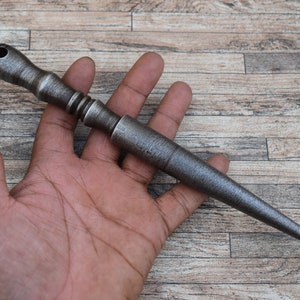 May include: A silver-colored, pointed, metal tool with a handle. The handle has a small hole and a decorative ring. The tool is held in a person's hand.