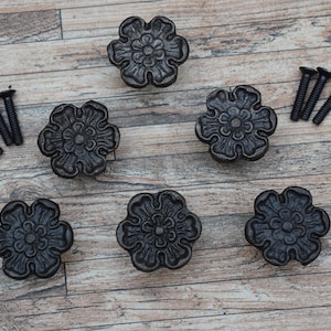 May include: Six black metal flower-shaped drawer pulls with intricate details. The pulls are arranged in a semi-circle on a wood surface. Four black screws are also included.