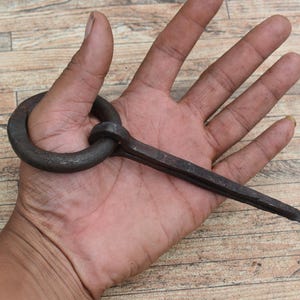 May include: A hand-forged, dark metal ring and spike tool. The ring is circular, and the spike is rectangular with a pointed end. The tool is held in a person's hand against a wooden background.