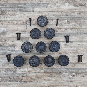 Op de afbeelding: Een set van twaalf zwarte metalen kastknoppen met een bloemmotief. De knoppen zijn in een driehoekige vorm op een houten oppervlak gerangschikt.