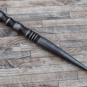 Puede incluir: Una herramienta larga, puntiaguda, de metal con un mango de madera. El mango tiene un agujero en la parte superior y está decorado con una serie de anillos.