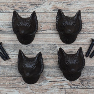Könnte beinhalten: Vier schwarze, detaillierte Schubladenknöpfe aus Gusseisen in Form eines Fuchs Kopfes. Die Knöpfe sind auf einem Holzgrund mit zwei schwarzen Schrauben abgebildet.