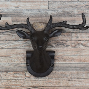 Könnte beinhalten: Ein schwarzer Wandhaken in Form eines Hirschkopfes aus Gusseisen mit detailliertem Geweih und einer Halterung. Der Hirschkopf ist an einer Wand aus Holzplanken befestigt. Der Haken ist zum Aufhängen von Gegenständen bestimmt.