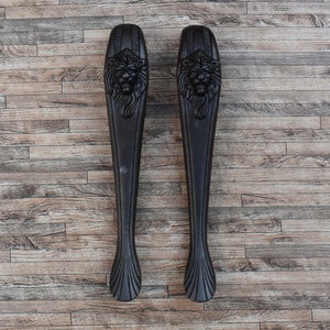 May include: Two black metal door handles with lion head designs. The handles are on a wooden background.