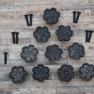 Op de afbeelding: Twaalf zwarte metalen bloemvormige ladegrepen met een gedetailleerd ontwerp, en acht zwarte metalen schroeven.