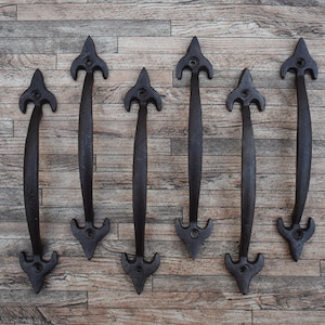 Könnte beinhalten: Sechs schwarze Türgriffe aus Gusseisen mit einem dekorativen pfeilartigen Design. Die Griffe sind auf einer Holzoberfläche angeordnet. Jeder Griff ist etwa 20 cm lang.