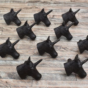 Op de afbeelding: Een verzameling van negen zwarte wandhaken in de vorm van een eenhoornkop, elk met gedetailleerde manen en een enkele spiraalvormige hoorn. Deze decoratieve hardware-onderdelen zijn in een rasterpatroon gerangschikt en hebben een matte afwerking.