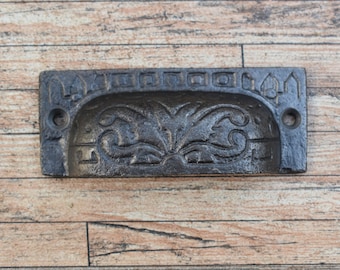 May include: A black cast iron drawer pull with an ornate floral design. The pull has a rectangular shape with rounded edges and two small holes for mounting.